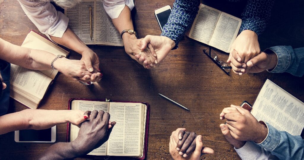 Six people in a prayer circle holding hands over a table with their bibles laying open.