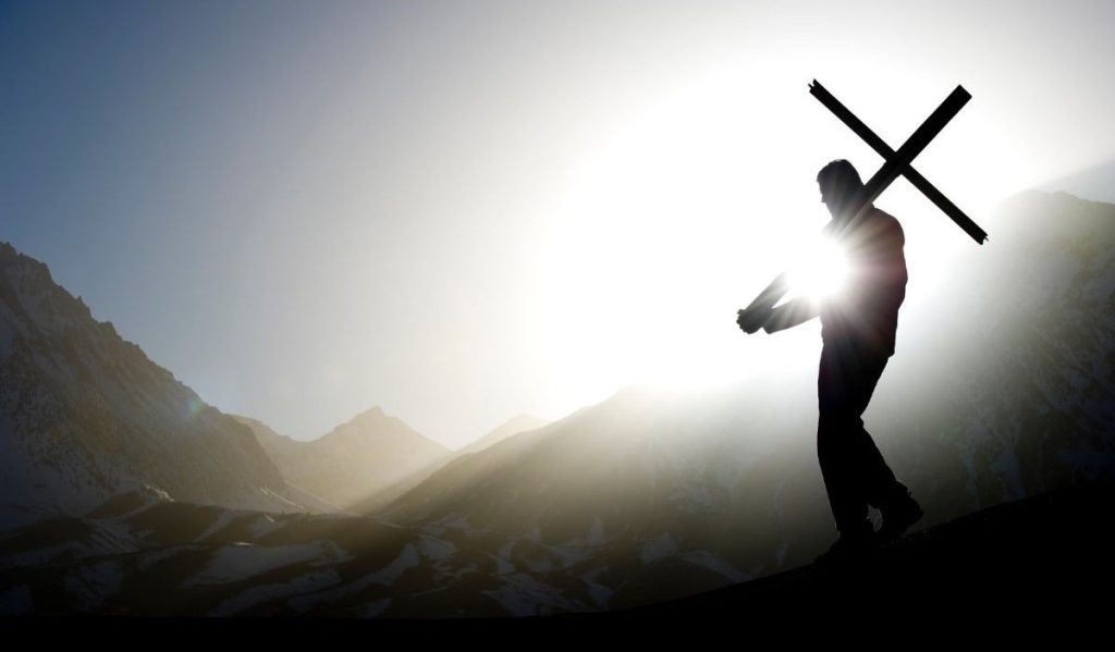 Image of a Man carrying a cross down a mountain with the sun rising in the background. 